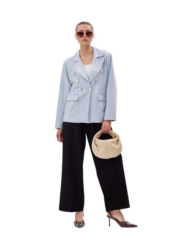 Woman wearing a light blue blazer and black pants, holding a beige handbag on a white background