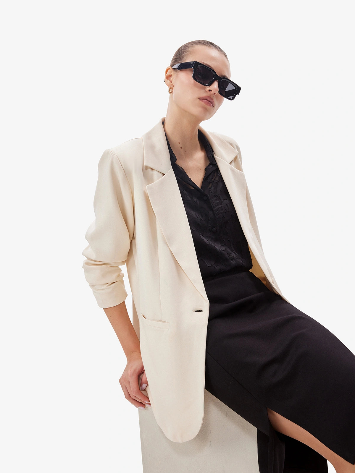 Woman wearing a beige blazer over a black dress with sunglasses on a white background